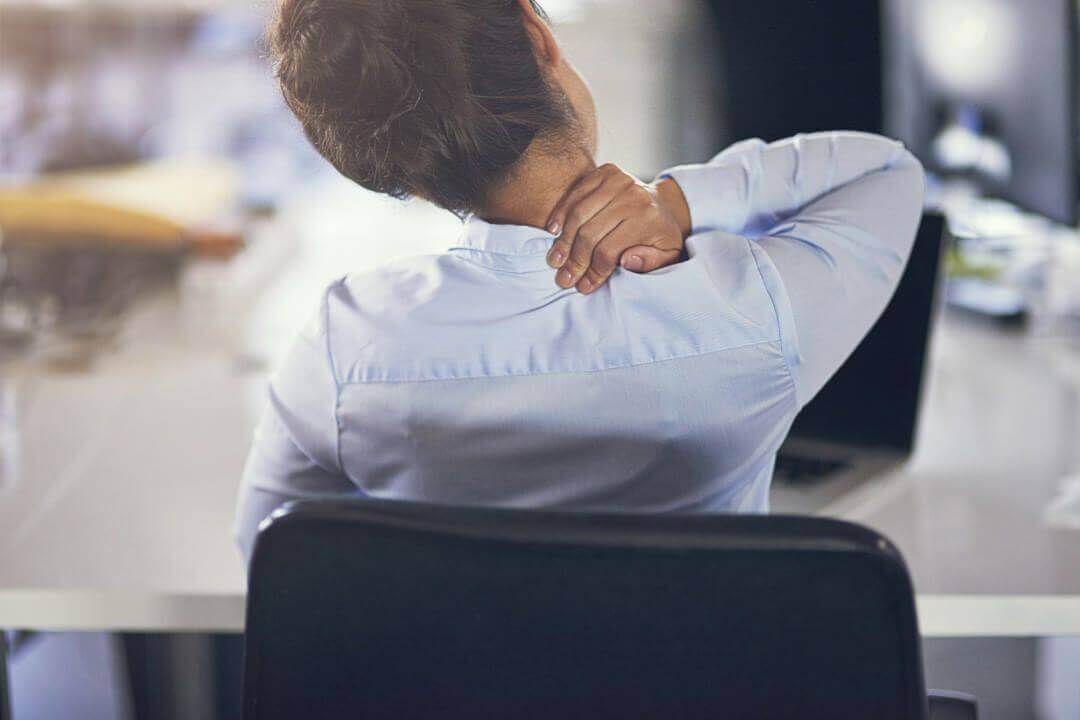 Office worker sitting at a desk with visible neck and shoulder strain at the end of a long workday